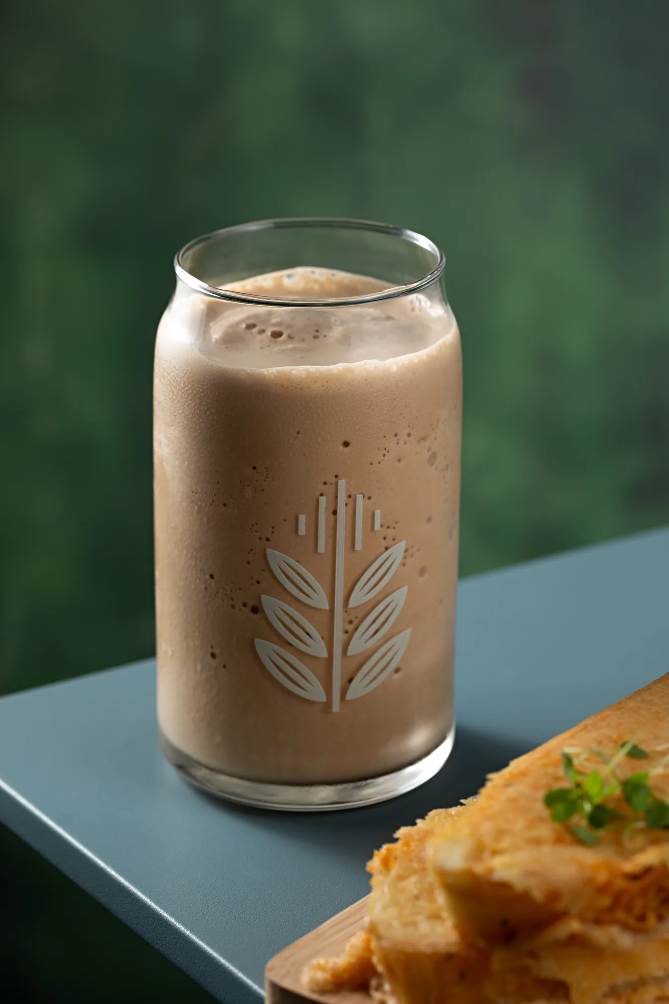 Copo de vidro com o símbolo da espiga da marca, cheio de café gelado e cremoso, sobre mesa azul, ao lado de uma fatia de focaccia tostada